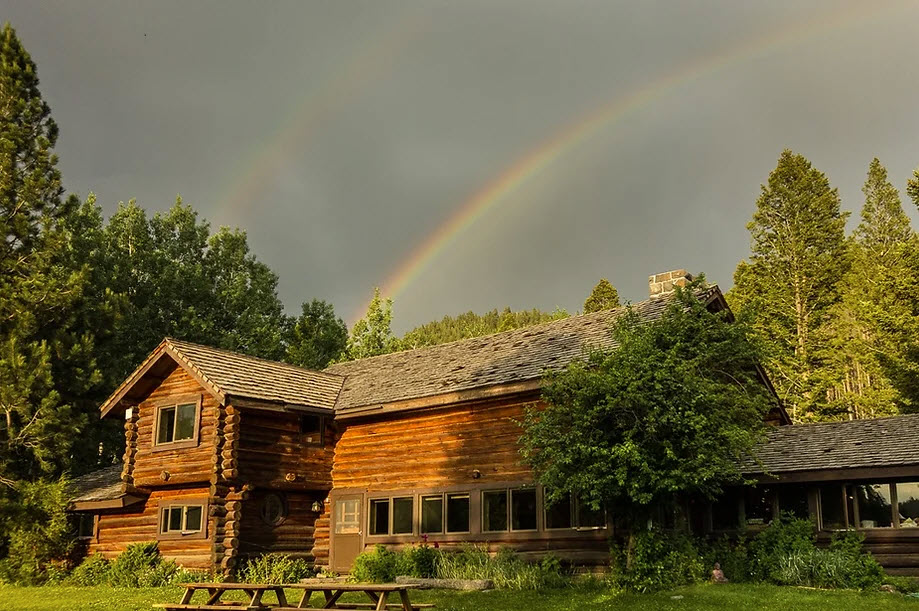 Double rainbow in sky above large log cabin surrounded by trees at Feathered Pipe Ranch in Montana.
