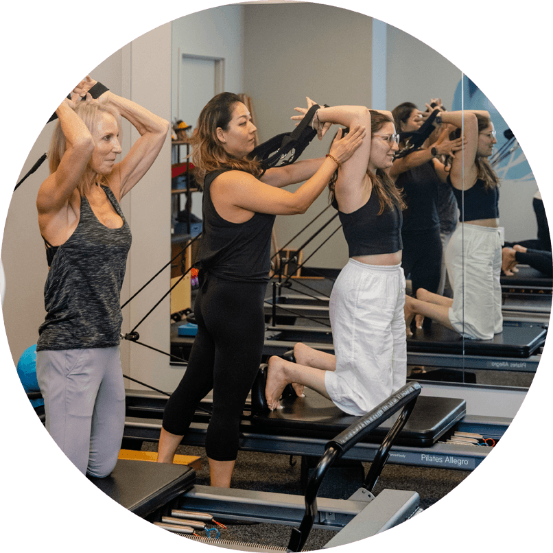 Pilates instructor guides students who are on knees with hands behind head gripping resistance bands.