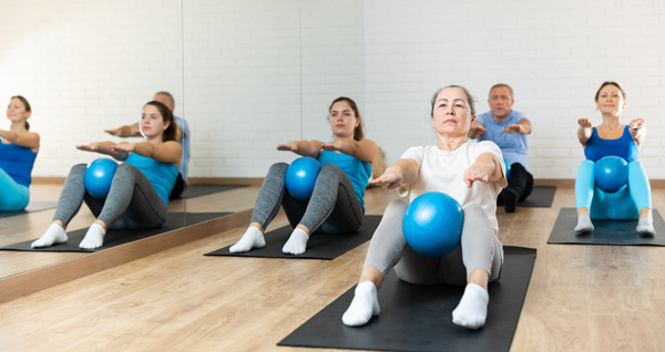 Elderly woman doing strength training mat pilates with ball in gym area.