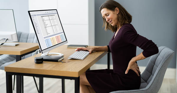 A women sitting in a chair at office desk with painful look on face while placing hand on lower back.