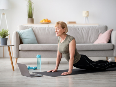 Mature women doing yoga pose on mat in her home watching a live streaming online tutorial on laptop.