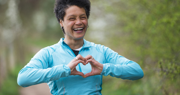 A mature black woman wearing sporty clothing smiling while making a heart shape with her hands.