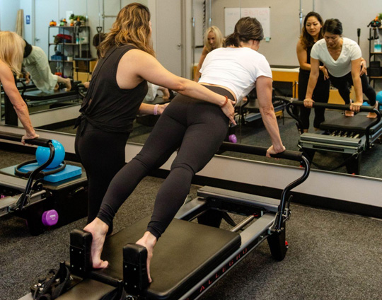 A Pilates instructor assisting a student on a Pilates Reformer machine to ensure proper positioning.