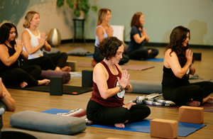 Group yoga session with students sitting barefoot on mats with hands together in yoga pose.